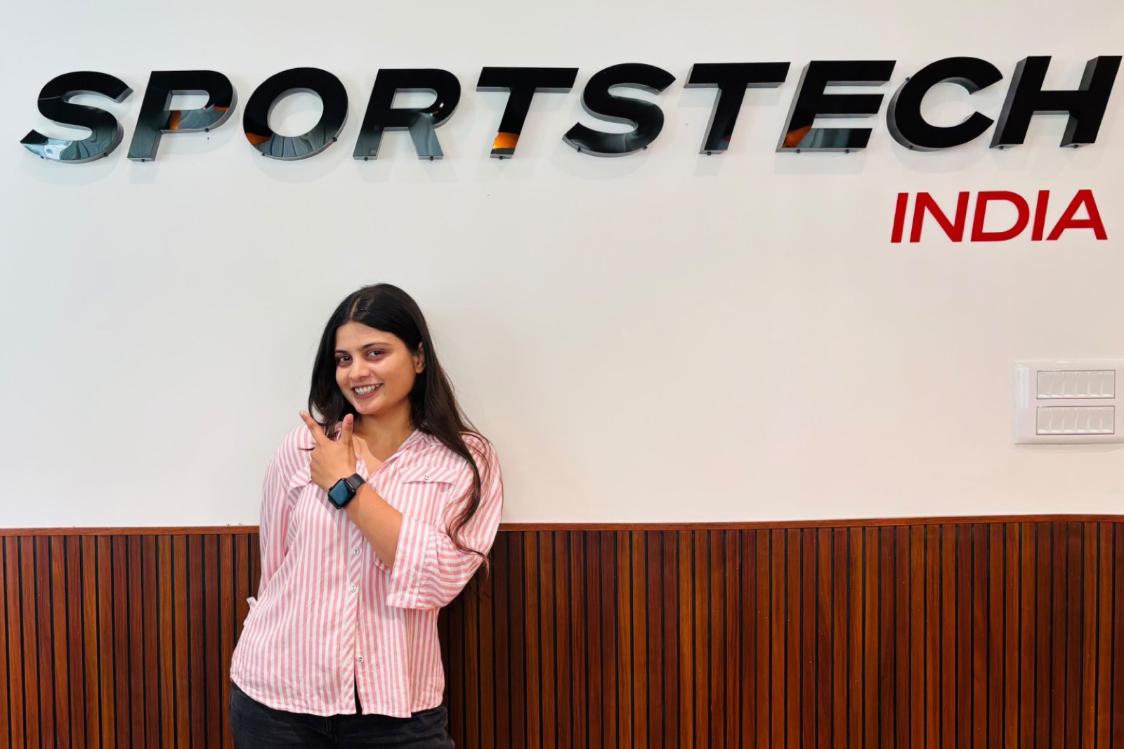 Urmi at the Sportstech India office in Bangalore, pointing up at the Sportstech sign